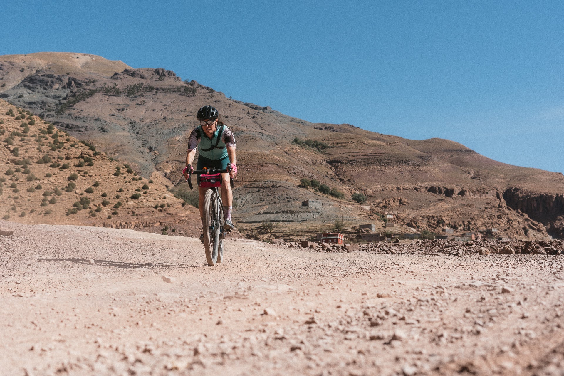 Cyclist riding a gravel bike on a rocky dirt road with mountainous terrain in the background under a clear blue sky.