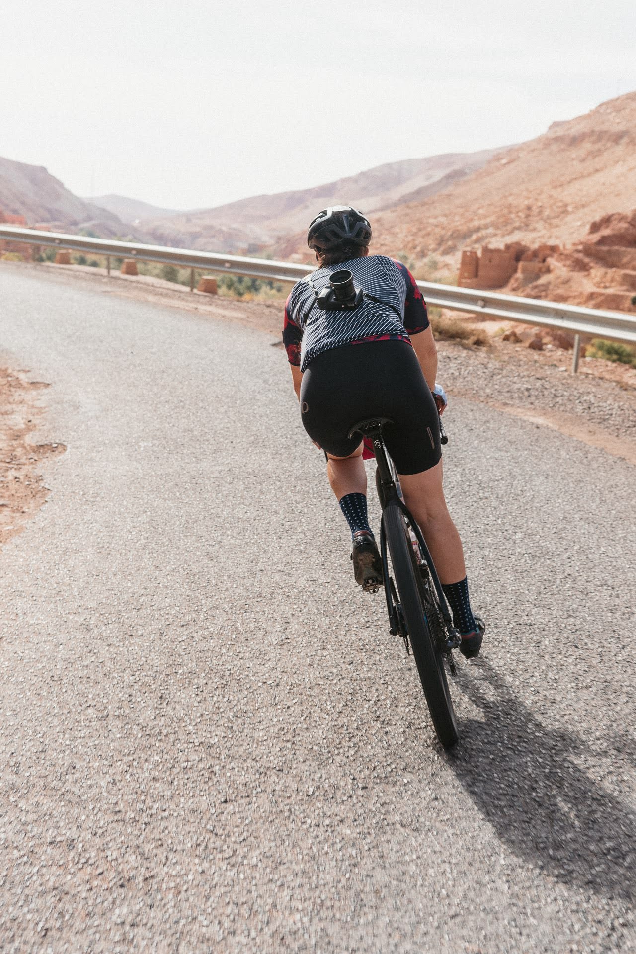 Cyclist wearing helmet and camera mounted on back riding on a paved mountain road with guardrail and barren hills.