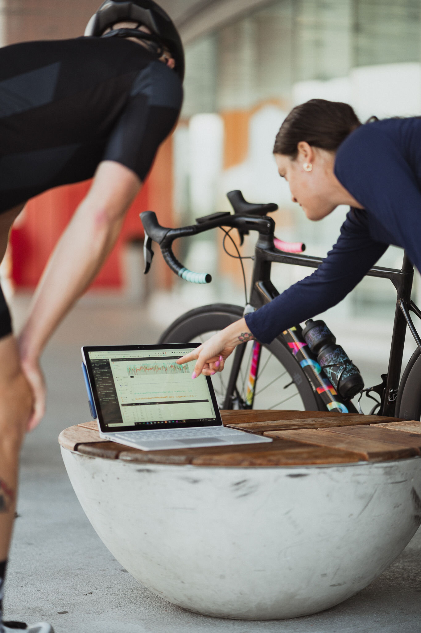 A woman points at a laptop screen showing data graphs while a man in cycling gear looks on beside a road bike.