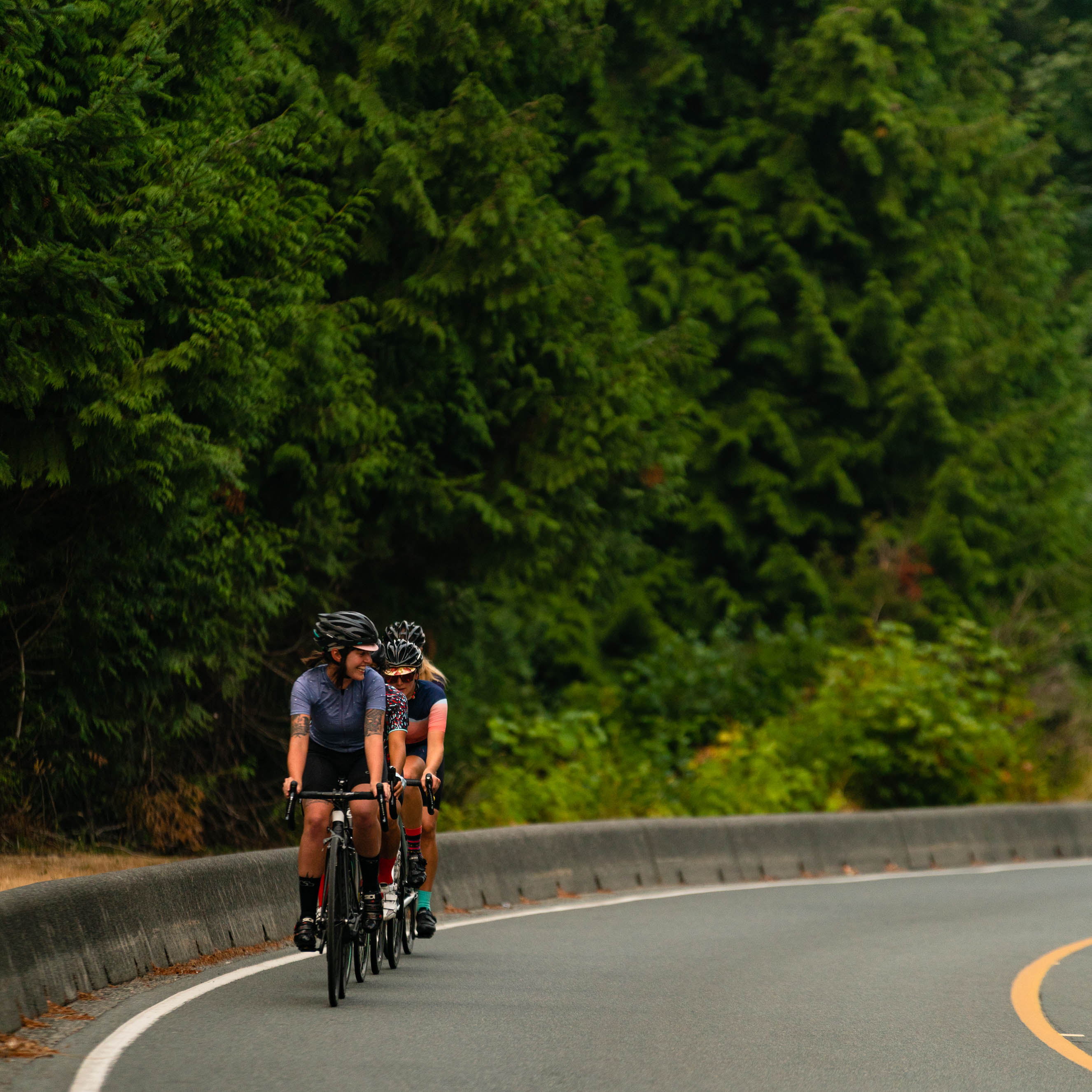 Two cyclists riding on a curved road alongside dense green foliage, both wearing helmets and cycling jerseys.