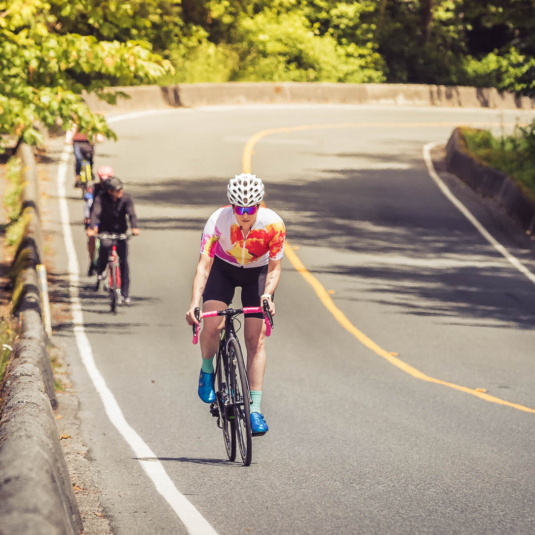 Cyclist wearing a white helmet and colorful jersey riding a bike uphill on a tree-lined road with other cyclists behind.